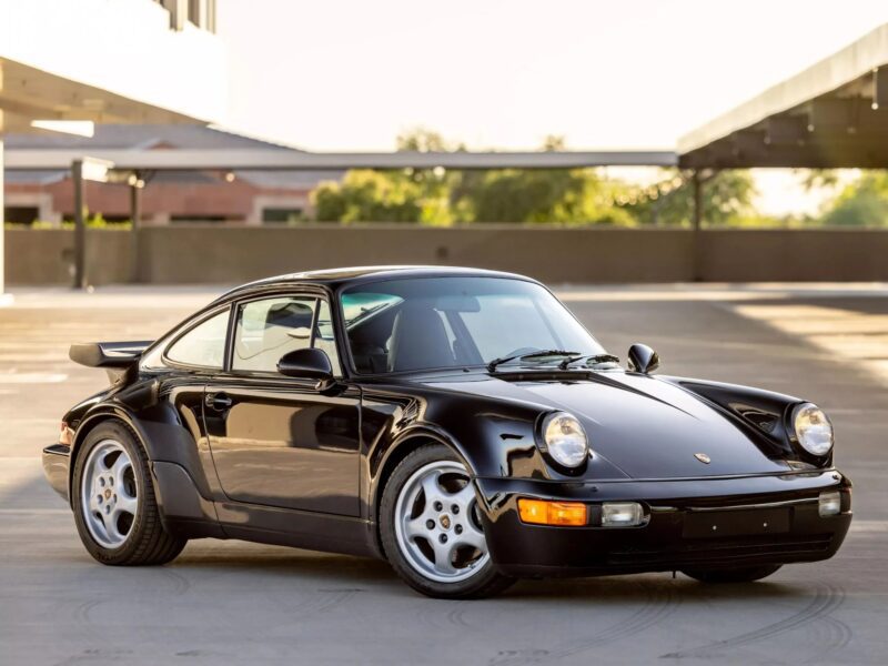 A black Porsche 911 sports car, one of the best air-cooled Porsches, is parked in an outdoor lot with modern buildings and trees in the background.