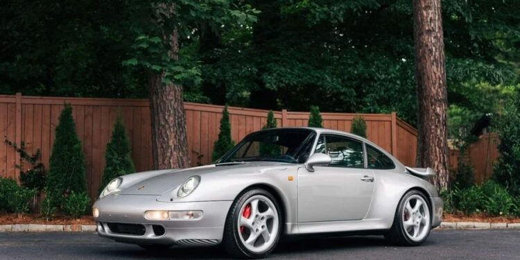 A silver Porsche 911, one of the Best Air-Cooled Porsches, is parked on a paved surface in front of a wooden fence and green trees.