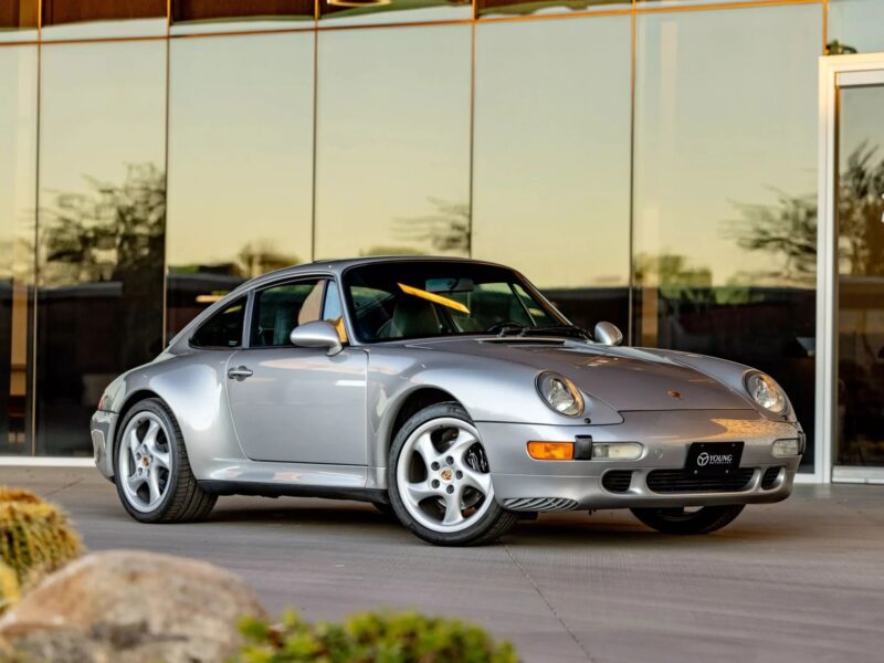 A silver Porsche coupe, one of the best air-cooled Porsches, is parked on a concrete surface in front of a modern glass building, viewed from the front left side.