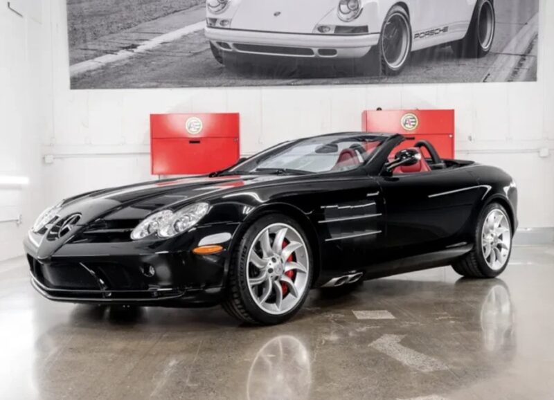 A luxurious black convertible sports car with a red interior is gracefully parked in a showroom, accompanied by a striking black and white photograph of a classic car. This Mercedes-Benz masterpiece is proudly on display and available for sale to discerning enthusiasts.