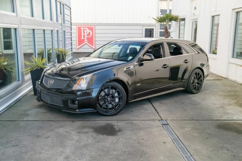 A luxurious black Cadillac CTS-V wagon, renowned among performance wagons, is parked on a concrete surface near a building with glass windows and potted plants.