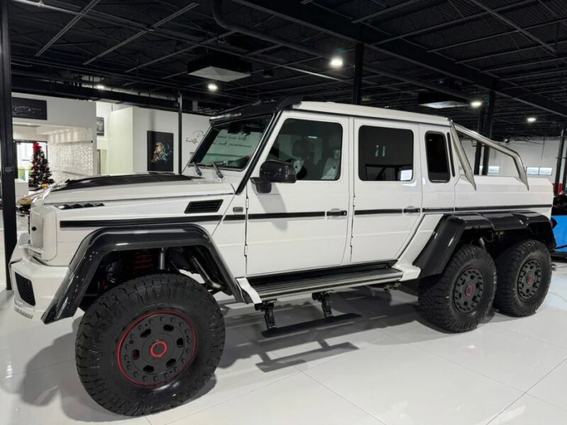 A luxurious white six-wheeled off-road vehicle with sleek black accents is proudly on display in the showroom, a testament to Mercedes-Benz's craftsmanship and now available for sale.