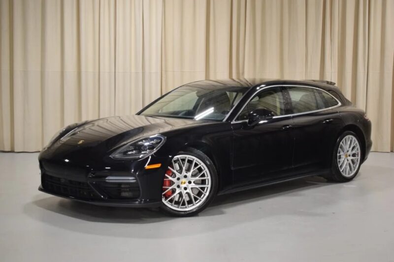 A black Porsche Panamera luxury performance wagon with silver alloy wheels is elegantly parked indoors on a light-colored floor, framed by beige curtains.