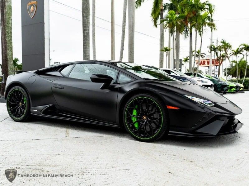 A black Lamborghini Huracan with green-accented wheels is parked outdoors near palm trees and a Lamborghini dealership sign, showcasing the sleek V10 supercar for sale.