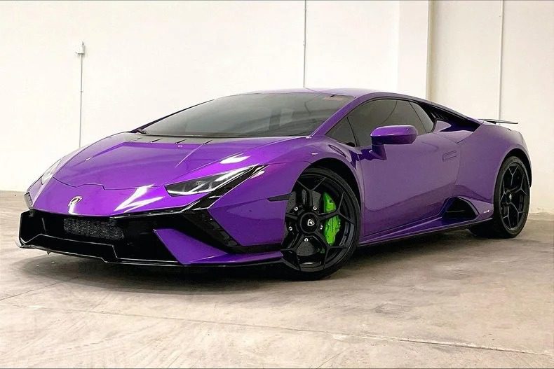 A striking purple Lamborghini Huracan Tecnica with green brake calipers is parked in a spacious garage, showcasing the allure of this V10 supercar. Now for sale, it's the perfect blend of power and style waiting for a new owner.