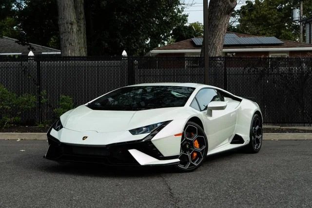 A pristine Lamborghini Huracan is parked on a street, its sleek lines contrasting against the black fence and trees in the background.