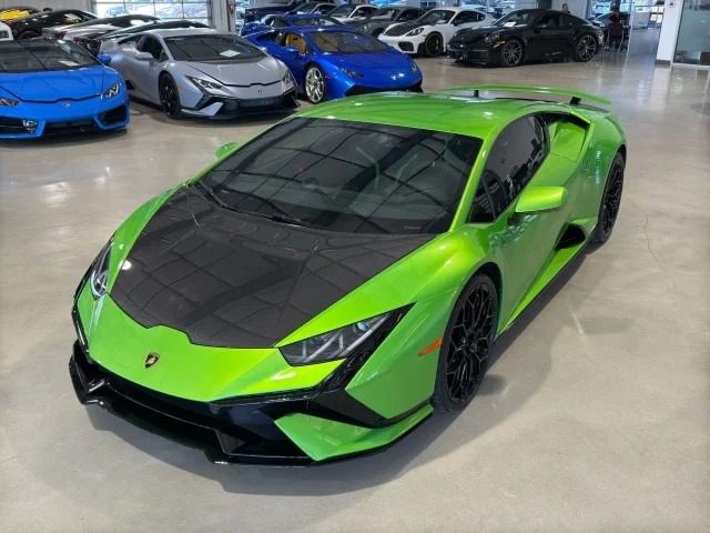 A green Lamborghini Huracan with a black hood is parked in a showroom, surrounded by various other sports cars. This V10 supercar, showcasing its stunning design and performance pedigree, catches the eye among the elite Tecnica for sale.