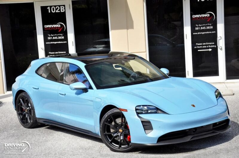 A light blue Porsche luxury performance wagon is parked outside a building, its sleek design perfectly framed by the glass doors and "Driving Emotions" signs.