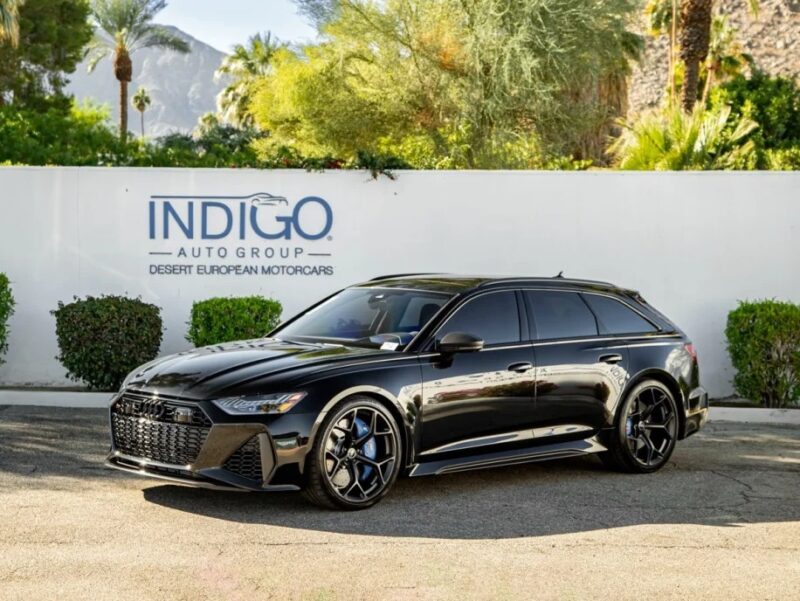 A black sports car is parked in front of a wall with "Indigo Auto Group Desert European Motorcars" signage. The backdrop, featuring trees and mountains, enhances the allure associated with luxury performance wagons.