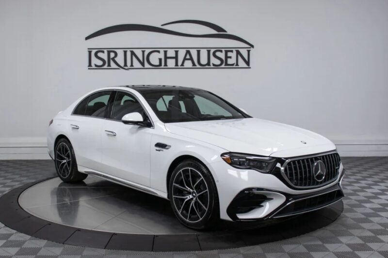 A luxury white Mercedes-Benz sedan is elegantly displayed indoors on a checkered floor, with the Isringhausen logo proudly showcased on the wall behind it, signaling that this exquisite vehicle is available for sale.