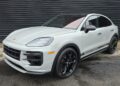 A white Porsche Cayenne Turbo GT, a true luxury performance SUV with black wheels and "Porsche" side decals, is parked on wet pavement in front of a dark corrugated metal wall.