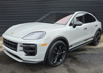 A white Porsche Cayenne Turbo GT, a true luxury performance SUV with black wheels and "Porsche" side decals, is parked on wet pavement in front of a dark corrugated metal wall.