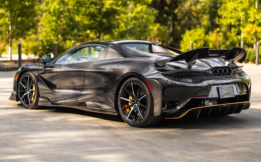 A sleek black sports car, reminiscent of the all-time greats like McLarens, is parked outdoors. It showcases its rear design and aerodynamic features with trees in the background, exuding an aura of luxury and expense.