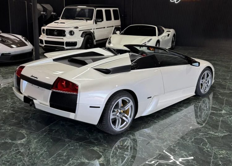 A pristine white convertible sports car, possibly a Lamborghini Murcielagos, is parked indoors on a gleaming marble floor. In the background, a sleek white SUV and another sports car hint at the luxurious surroundings of Sant'Agata.