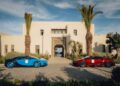 Two luxury hypercars, one blue and one red, elegantly parked in front of a grand modern beige building with palm trees and a clear sky.