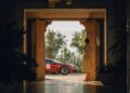 A person stands in an ornate Moroccan doorway, gazing at a stunning red Bugatti parked outside. Lush greenery and majestic mountains serve as the breathtaking backdrop.