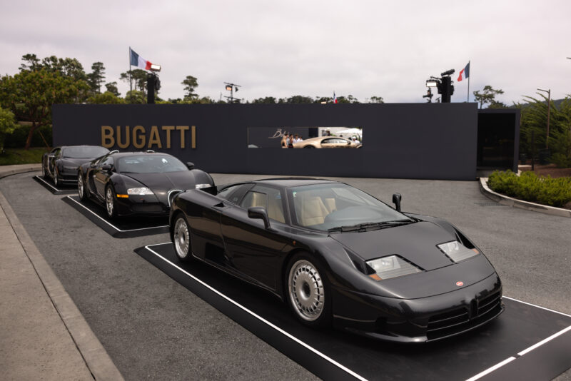 Three black Bugatti cars are displayed in a row outdoors at the Pebble Beach Concours d'Elegance, with a Bugatti sign and backdrop featuring a classic car image in the background.