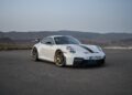 A white 2025 Porsche 911 GT3 with gold wheels is parked on a paved surface against a backdrop of mountains under a clear sky.