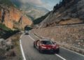 Two high-performance sports cars, including a sleek Bugatti hypercar, navigate a winding mountain road with rocky cliffs and a cloudy sky in the background.