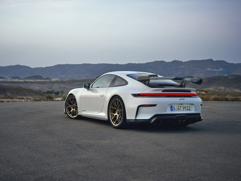 A white Porsche 911 GT3, featuring a Touring Package, is parked on a paved surface against the backdrop of mountainous terrain.