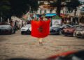 A person proudly holds a large Moroccan flag on a bustling street, where the roar of a Bugatti hypercar might easily harmonize with the vibrant atmosphere, amid parked cars and towering buildings in the background.