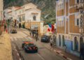 Three hypercars, including a sleek Bugatti, race through a narrow street lined with colorful buildings and animated crowds. French and Moroccan flags flutter above, set against a backdrop of rugged hills in this lively Moroccan scene.