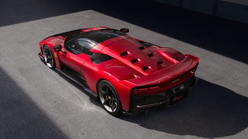 A red Ferrari sports car with a sleek design is parked on a sunlit asphalt surface, viewed from the rear three-quarter angle.