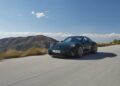 A dark green Porsche 2025 911 GT3 effortlessly cruises along a scenic road, its Touring Package adding a touch of elegance, with majestic mountains and a brilliant blue sky as the perfect backdrop.