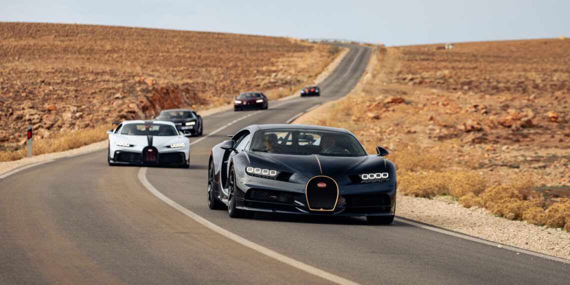 A line of luxury sports cars, including a sleek Bugatti hypercar, cruises along a rural road amidst the stunning desert landscape of Morocco.