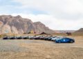 A row of hypercars, including a Bugatti, is parked in a Moroccan desert landscape with rocky hills in the background. Two camels and a small tent are visible in the distance.