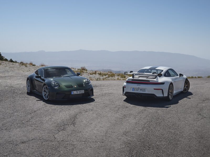 Two sleek sports cars, including a 2025 Porsche 911 GT3 with the Touring Package, are parked on a mountain road, offering a breathtaking view of distant hills under the clear blue sky.