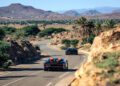 Two sleek sports cars, one a Bugatti hypercar, navigate the winding mountain roads of Morocco, their engines roaring against rocky terrain and distant hills under a clear sky.