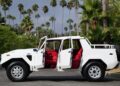 A striking white SUV, resembling a 1990 Lamborghini LM002, with its four doors open and vibrant red interior, is majestically parked on a street lined with swaying palm trees.