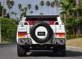 A white SUV with a spare tire on the back reads "LM002" on the rear plate. Palm trees and greenery line the street, showcasing an iconic 1990 Lamborghini LM002 cruising in style.