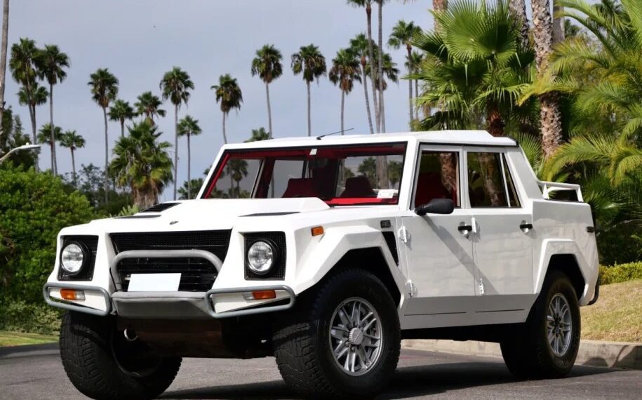 A 1990 Lamborghini LM002, an iconic white off-road vehicle, is parked on a street, surrounded by swaying palm trees and lush greenery. This rare Lamborghini for sale brings adventurous spirit and luxury to any terrain.