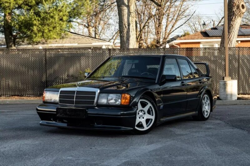 A nostalgic black vintage Mercedes-Benz sedan, reminiscent of classic 90s cars, is parked on a street, showcasing a prominent rear spoiler and sporty wheels. The background features trees and a fence.