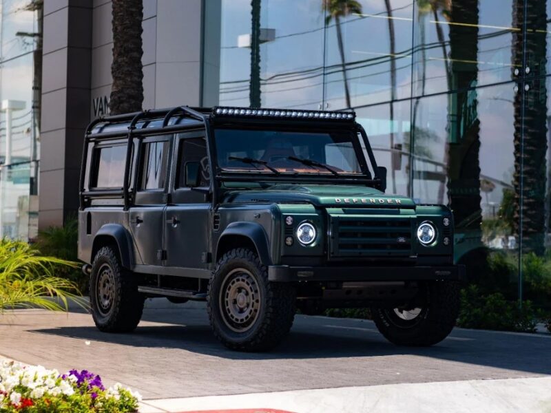 A black off-road vehicle reminiscent of 90s cars is parked on the street, with palm trees reflecting nostalgically in the nearby glass building.