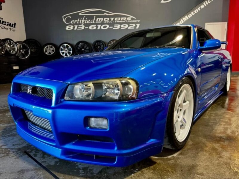 A bright blue sports car with white wheels is parked in a showroom, evoking nostalgia for 90s cars, surrounded by other tires and rims.