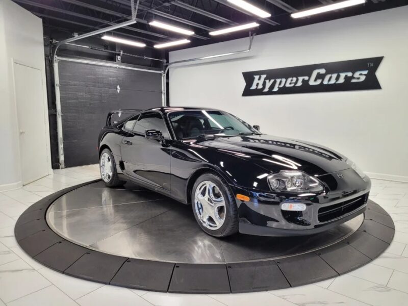 A black sports car dazzles on a rotating platform in the showroom, with "HyperCars" signage glowing in the background. Its sleek design evokes nostalgia for 90s cars, seamlessly blending classic charm with modern allure.