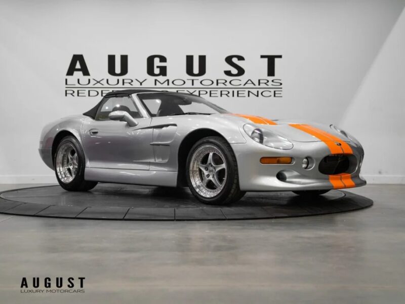 A silver sports car with orange stripes, evoking 90s nostalgia, is parked indoors against the backdrop of "August Luxury Motorcars" signage.