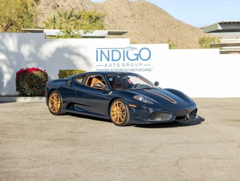 A striking blue Ferrari 430 Scuderia with gold rims sits in front of a wall emblazoned with "INDIGO AUTO GROUP," set against mountains and greenery. This prancing horse is for sale, offering the ultimate blend of performance and elegance.