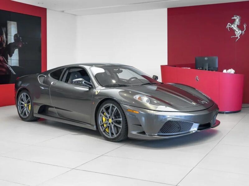 A gray Ferrari 430 Scuderia is showcased in a showroom with red and white walls. The sports car, adorned with yellow brake calipers and a prominent side vent, features the iconic Prancing Horse emblem, exuding elegance and performance.