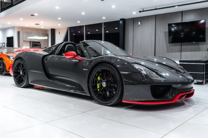 A sleek black Porsche 918 Spyder with red accents is parked indoors. This hypercar features an open top and polished alloy wheels, epitomizing luxury and performance.