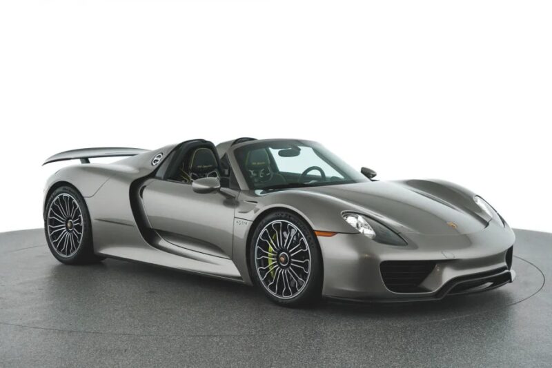 A sleek, silver Porsche 918 Spyder with an open-top design, large wheels, and a prominent rear spoiler rests elegantly indoors on a circular platform. This remarkable hypercar invites enthusiasts to experience its exhilarating performance and timeless design.