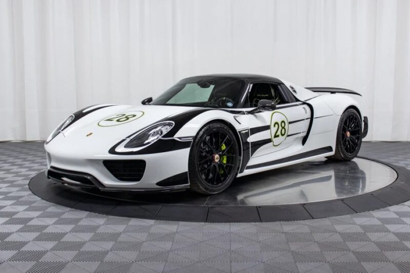 A sleek white and black Porsche 918 Spyder hypercar, proudly featuring the number 28 on its door, is displayed on a circular platform against a pristine white curtain backdrop.