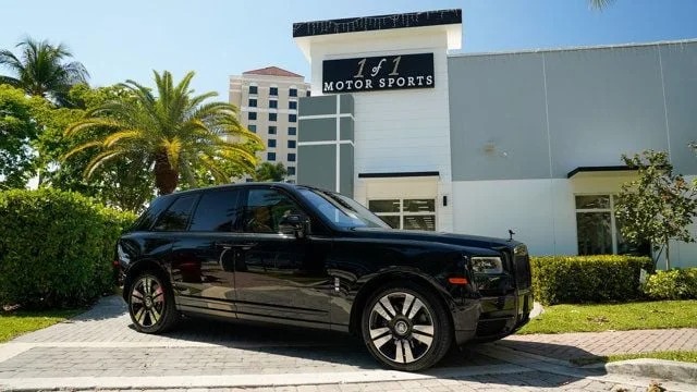 A Rolls-Royce Cullinan, the pinnacle of luxury SUVs, shines majestically in front of the "1 of 1 Motor Sports" building. With towering palm trees and an impressive multi-story backdrop, this scene exudes opulence and exclusivity.