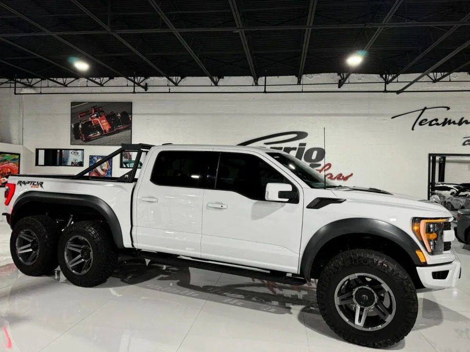 A striking white modified 6x6 truck is parked indoors, surrounded by automotive decor and displays. This stunning vehicle is sure to turn heads everywhere with its impressive design and powerful presence.