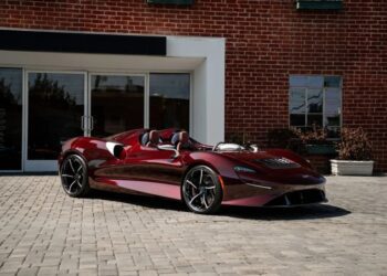A sleek, maroon McLaren Elva, the ultimate thrill machine, is parked on a brick driveway in front of a red brick building.