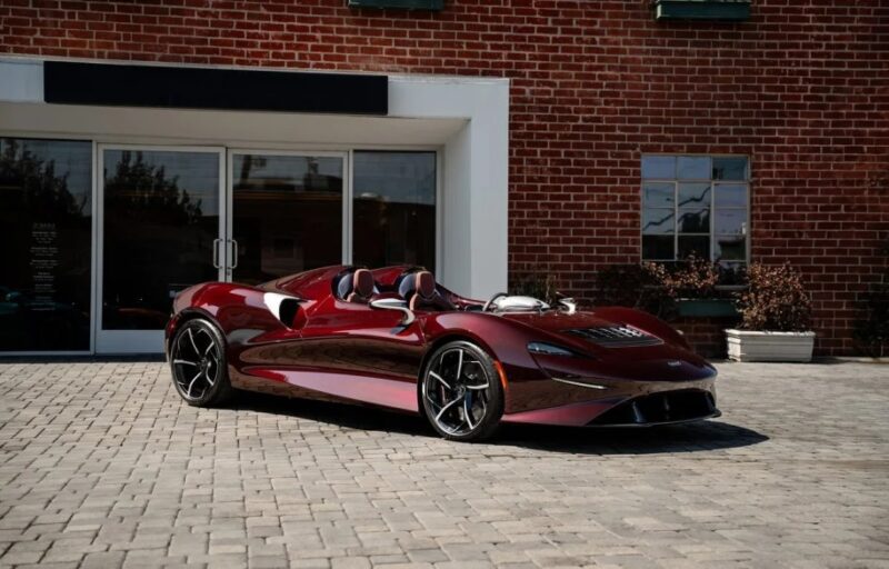 A sleek, maroon McLaren Elva, the ultimate thrill machine, is parked on a brick driveway in front of a red brick building.