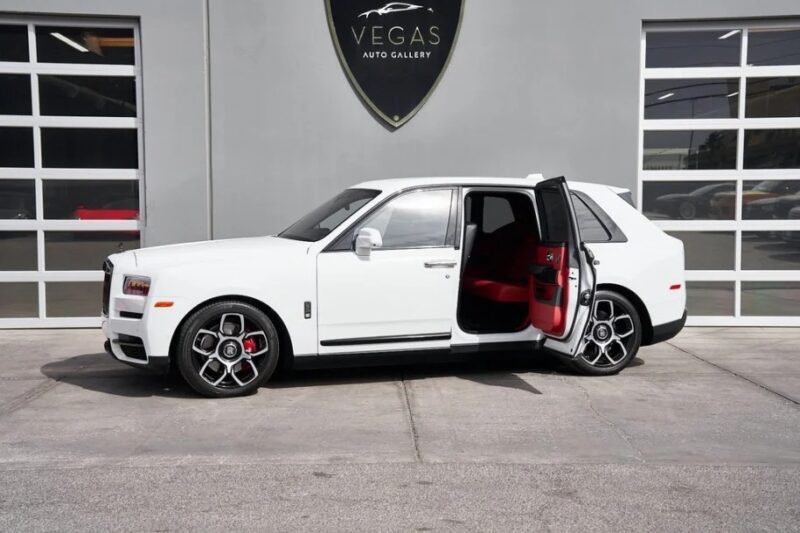 A white Rolls-Royce Cullinan with a rear passenger door open is parked in front of a building bearing the "Vegas Auto Gallery" sign. The red interior, epitomizing pinnacle luxury, is visible through the open door.